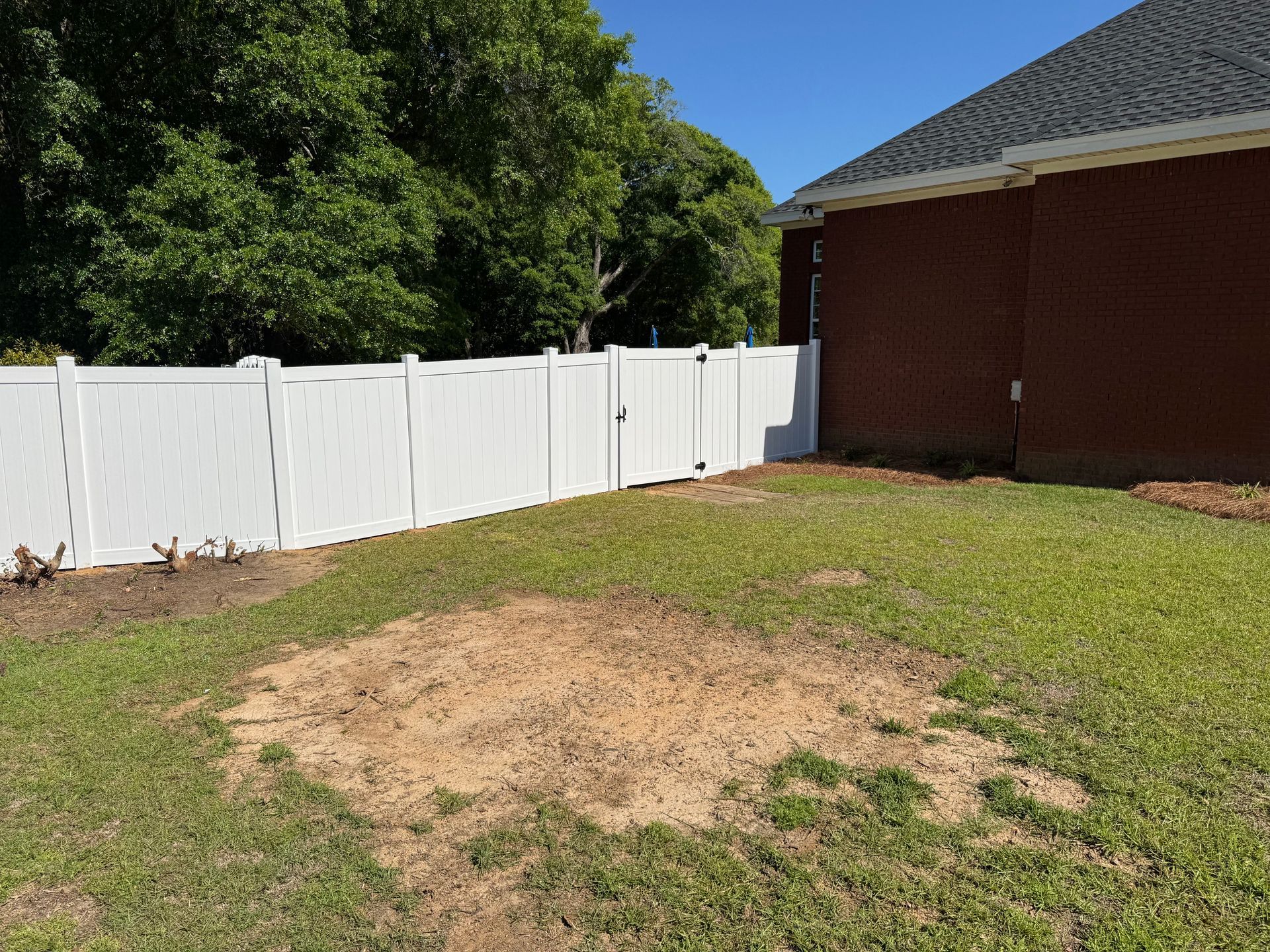 White fence in a backyard with a patch of dirt and brick house on a sunny day.