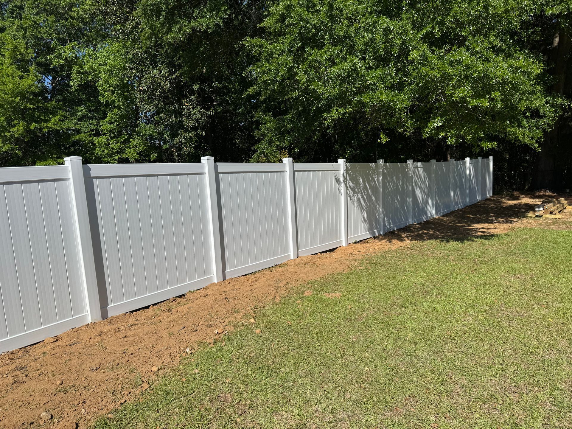 White vinyl lattice fence bordering grass and dirt with trees in the background.