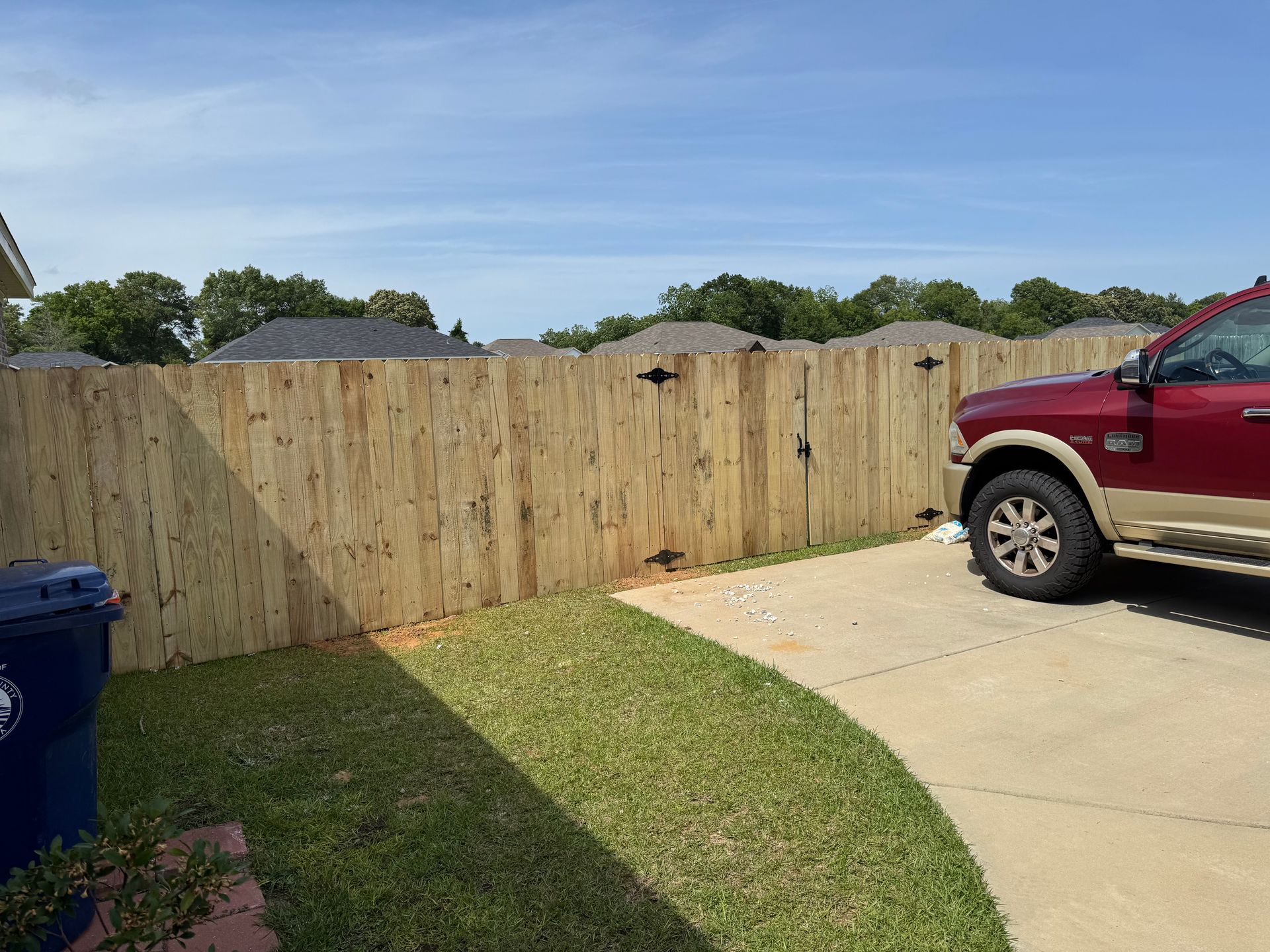 Wooden fence with a gate, next to a red pickup truck parked on a driveway, with green grass in the foreground.