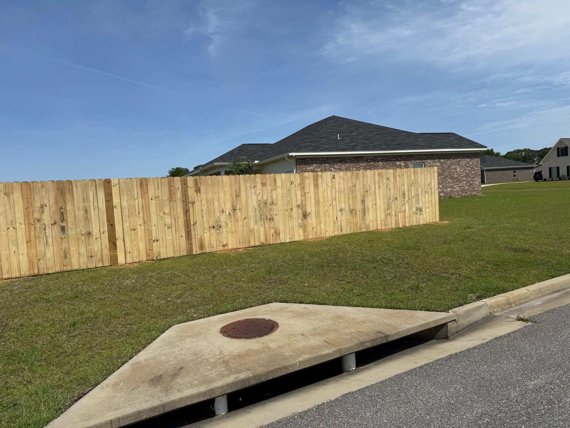 A wooden fence along a grassy lawn, with a house in the background under a blue sky.