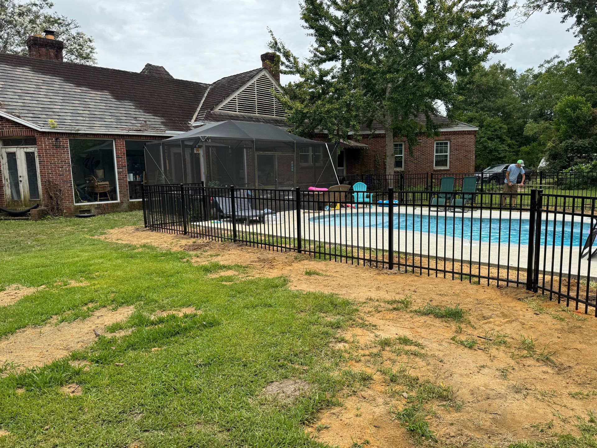 Backyard with pool, black fence, screened gazebo, and brick house with damaged roof.