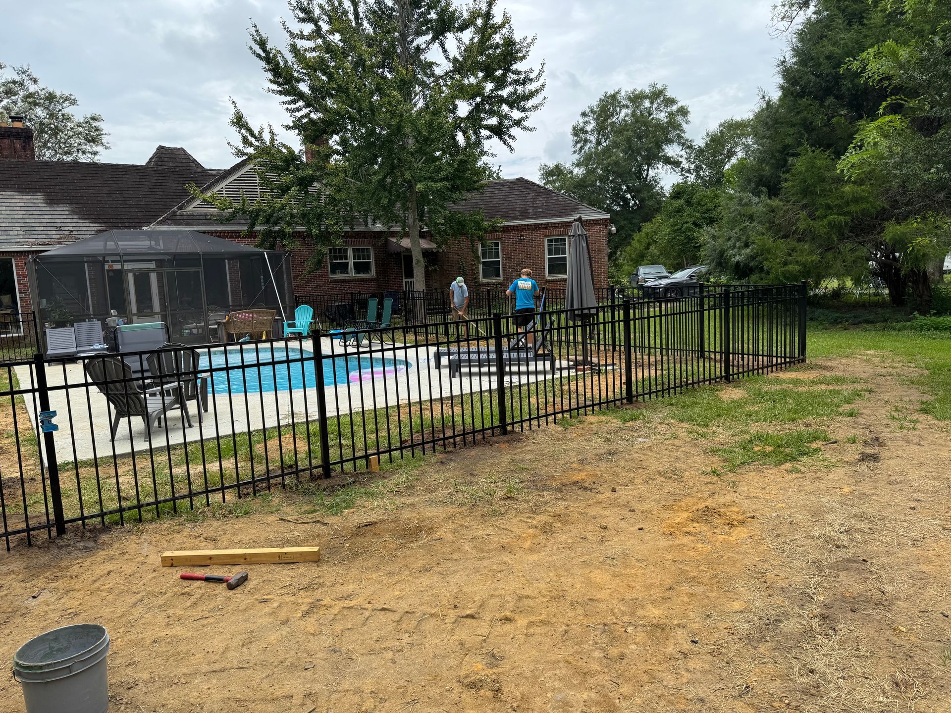 A black metal fence surrounds a backyard pool area, with a house and trees in the background.
