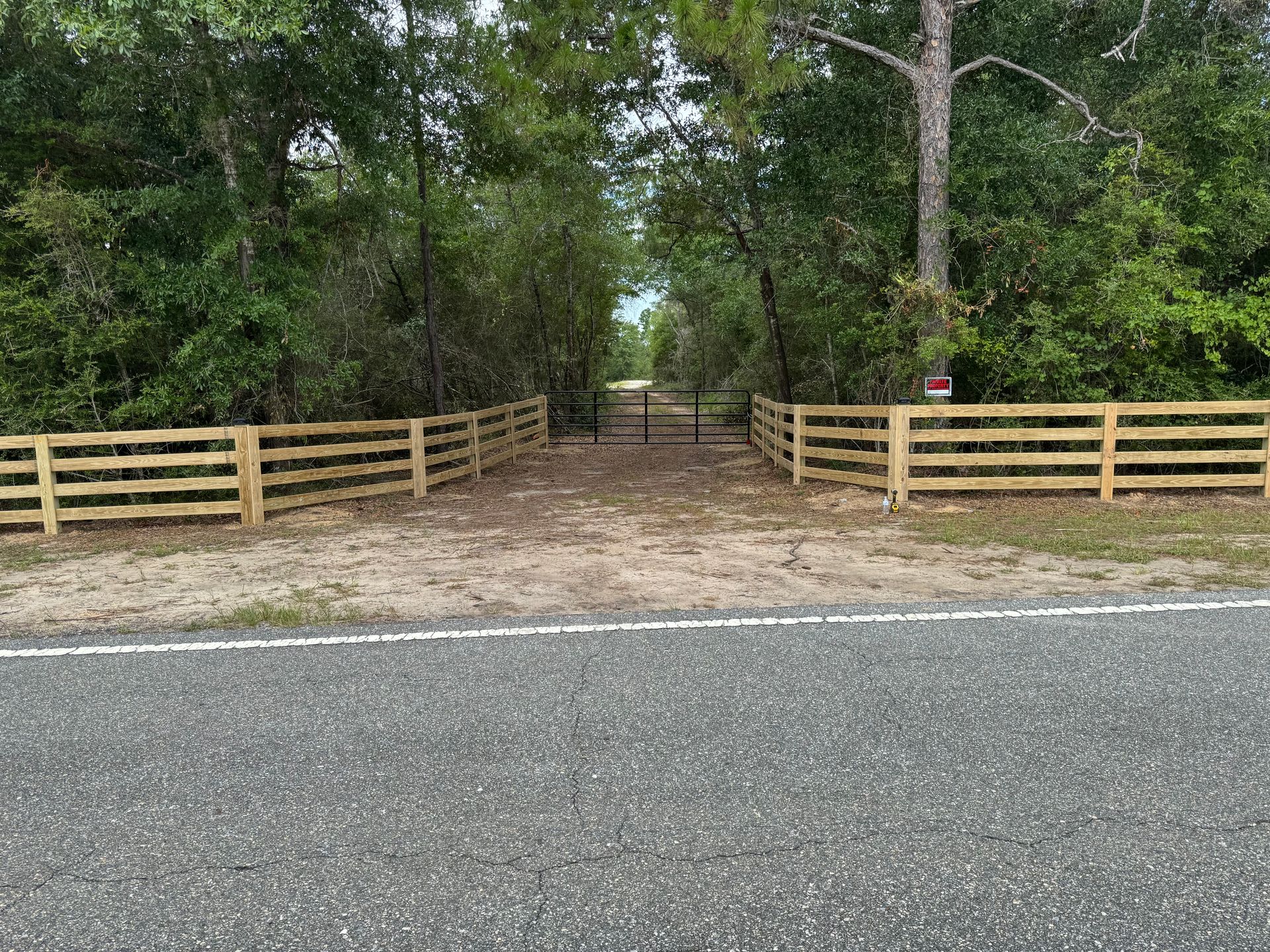 Wooden fence lines a gravel driveway leading to a gate, flanked by trees.