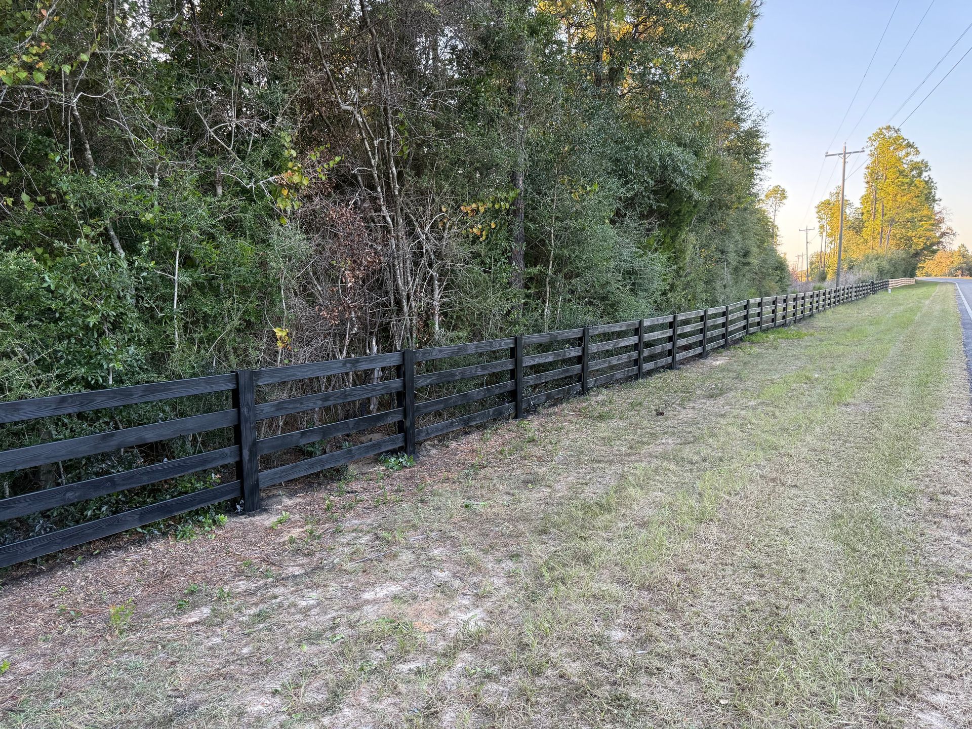 Black wooden fence bordering a roadside with green grass, trees, and blue sky.