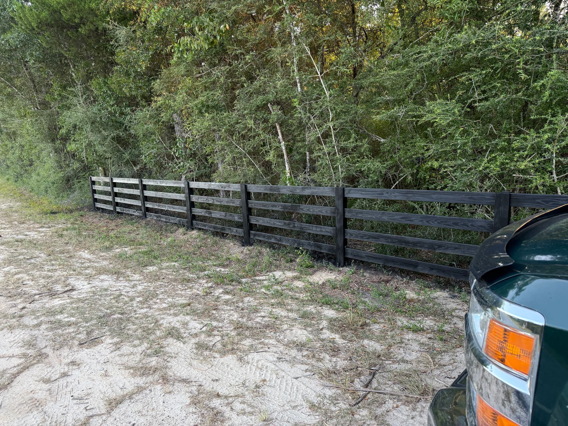 Black wooden fence along a sandy path with green foliage in the background, vehicle in foreground.