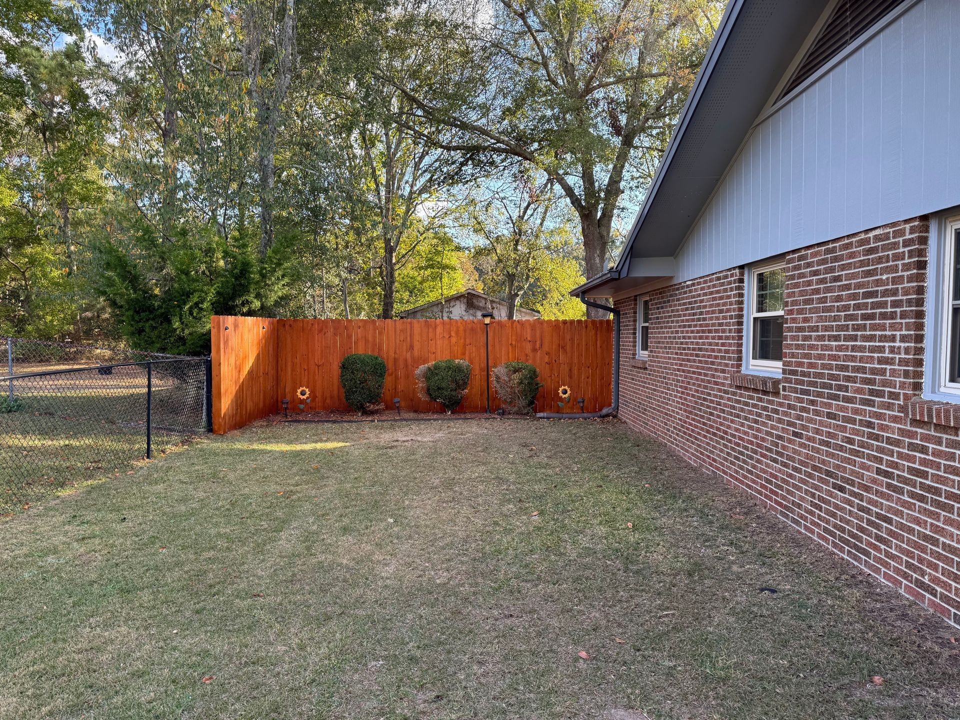 Backyard with a brown fence, brick house, green grass, and small bushes.