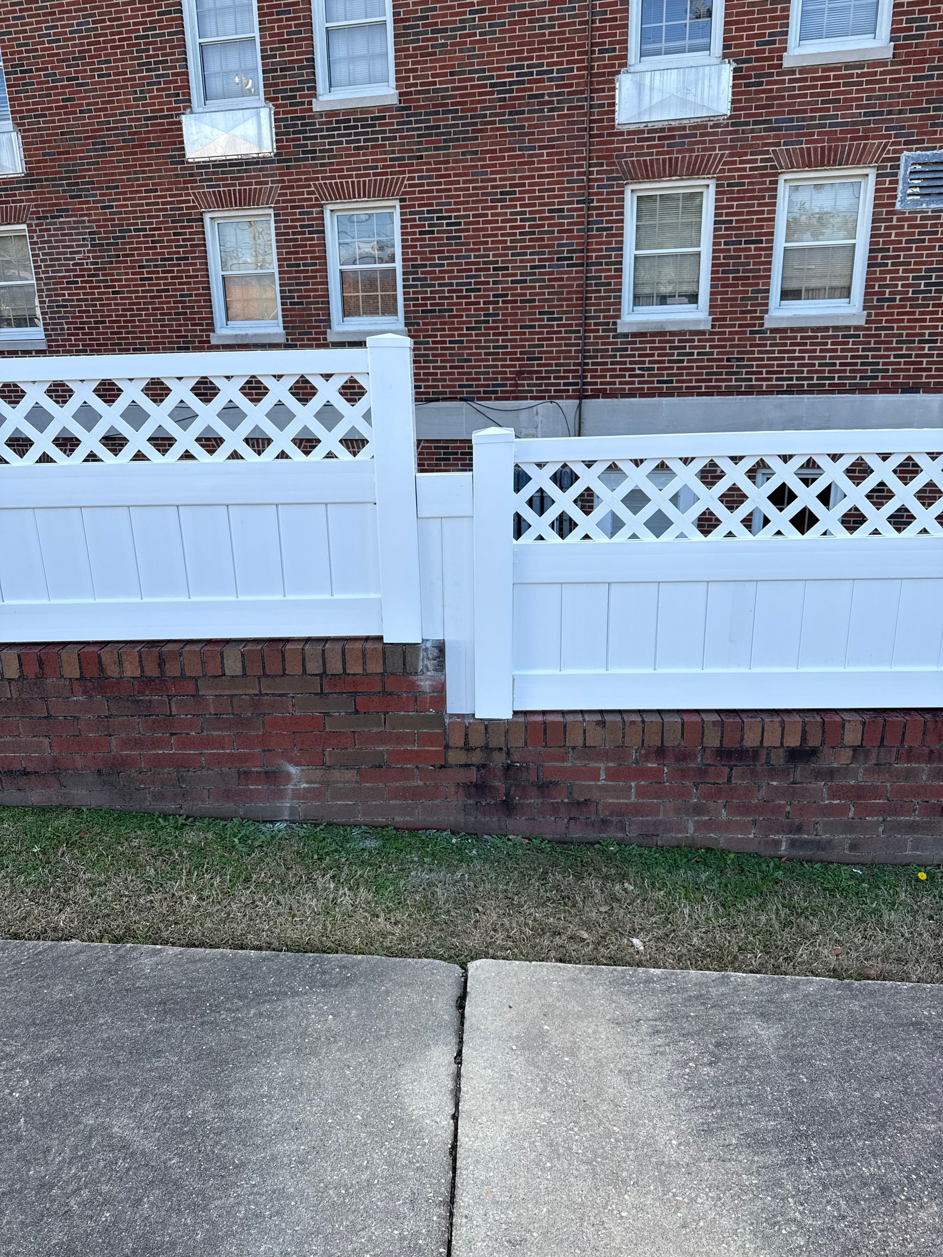 White lattice fence atop a brick wall in front of a brick building with windows.