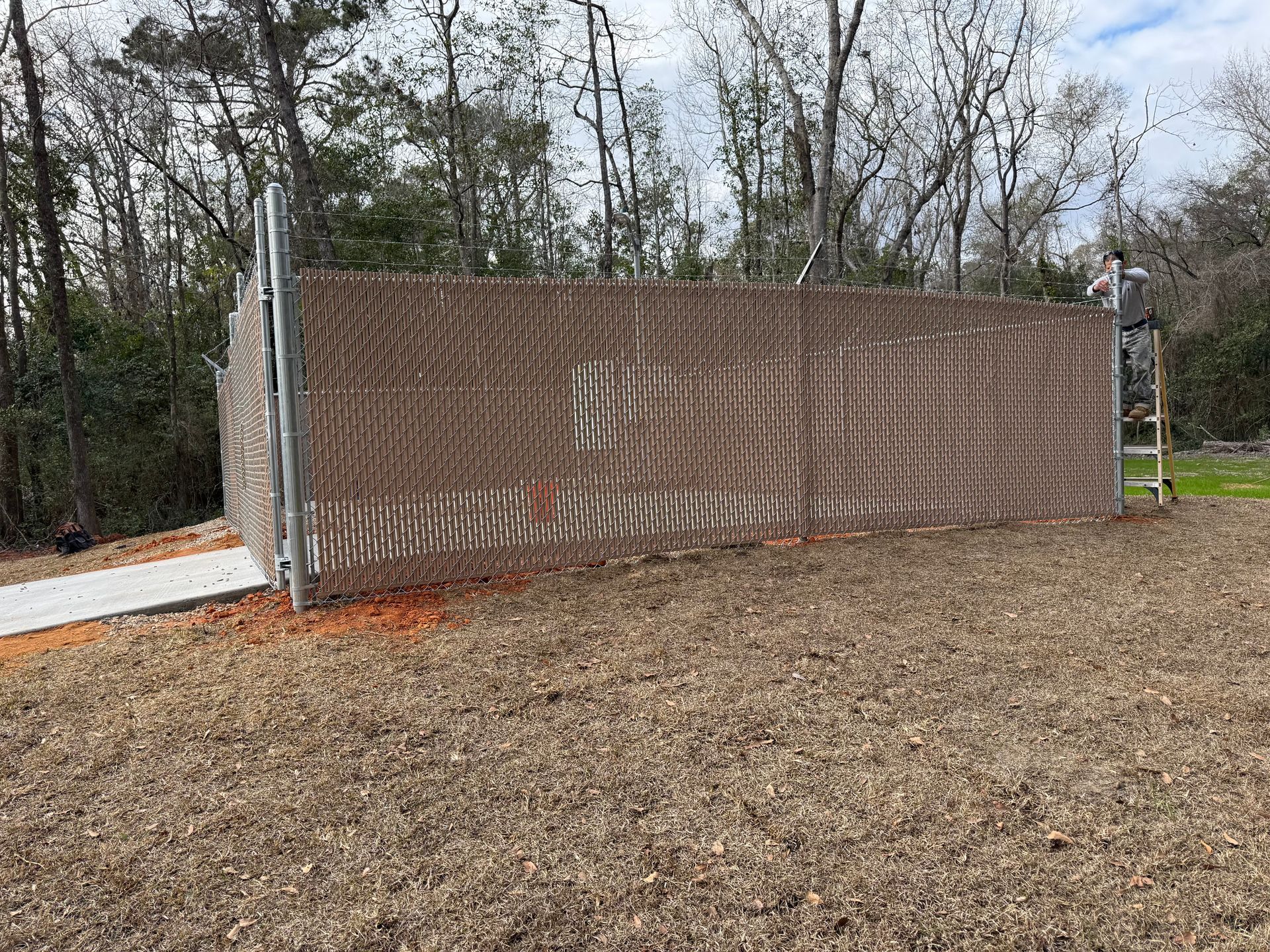 A woven artwork fence, brown and beige, stands in a grassy area with trees. A person on a ladder.