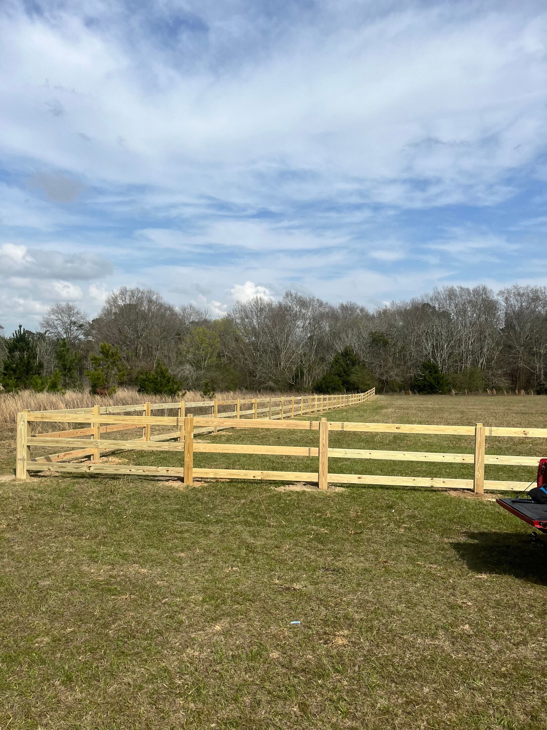 Wooden split-rail fence in a grassy field, under a cloudy blue sky. Trees in the background.