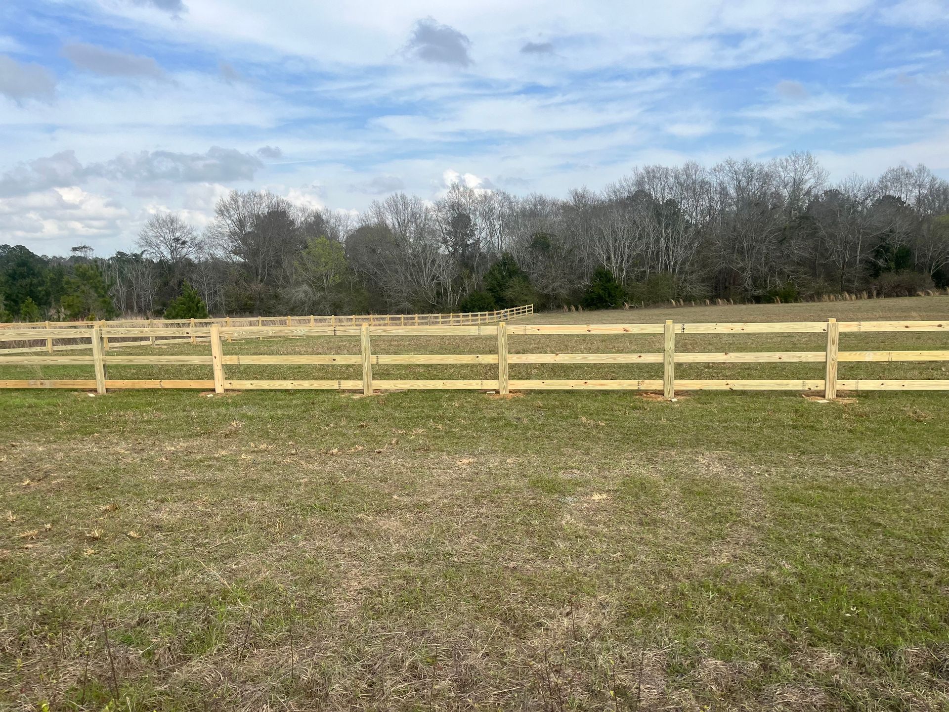 Wooden fence in a field with trees and a cloudy sky in the background.