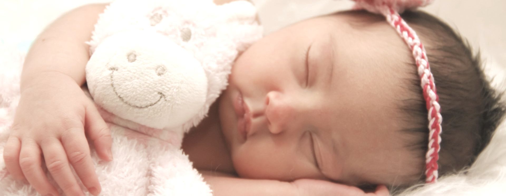 Sleeping baby cuddling a white teddy bear, wearing a pink headband on a soft bed