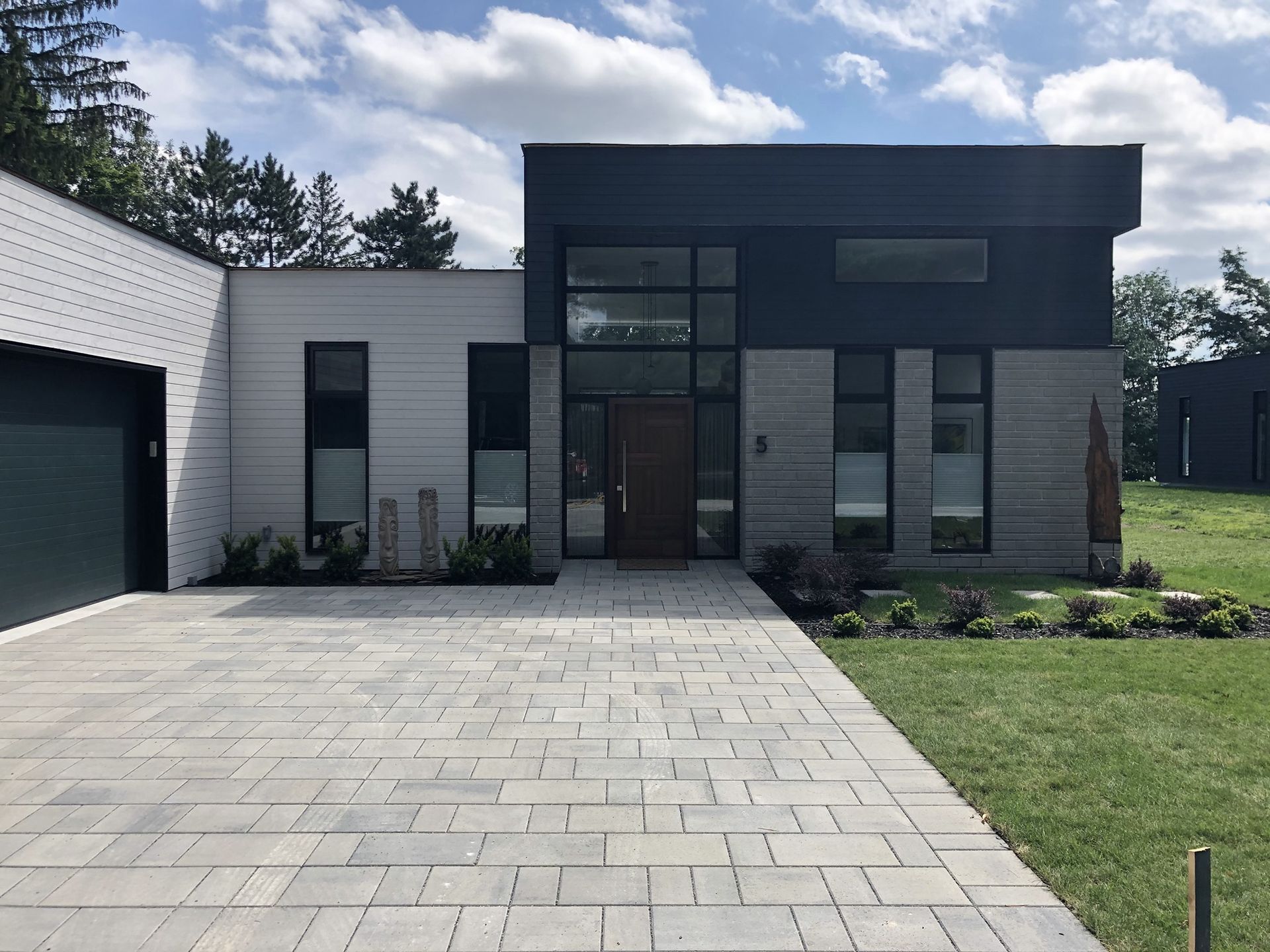 Une maison moderne de plain-pied avec une façade en pierre et en métal foncé, de grandes fenêtres, une porte d'entrée en bois et une allée pavée.
