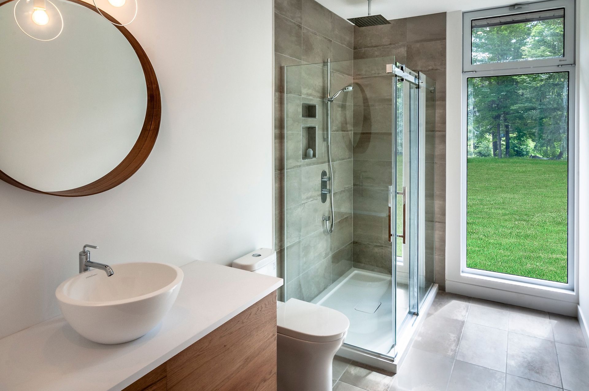Salle de bains moderne avec miroir rond, lavabo blanc, douche carrelée et fenêtre donnant sur une pelouse verdoyante.