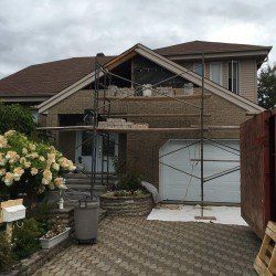 Maison en construction ; maçonnerie, échafaudage, toit brun, ciel gris nuageux.