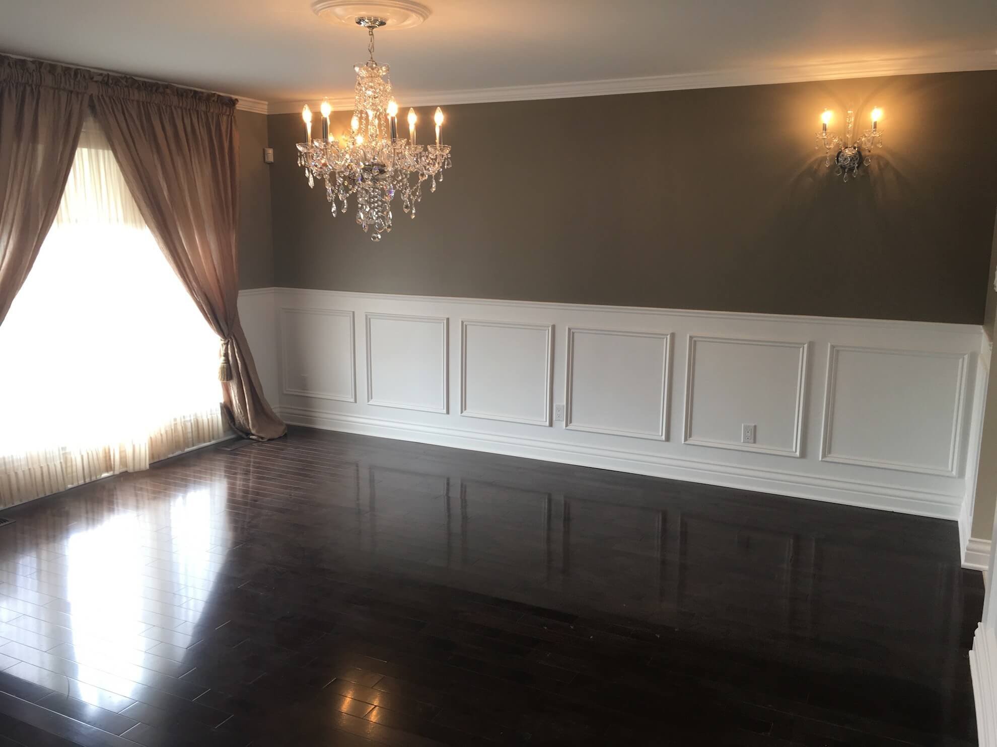Une salle à manger avec parquet foncé, murs taupe ornés de lambris blancs, un lustre en cristal et des rideaux drapés.