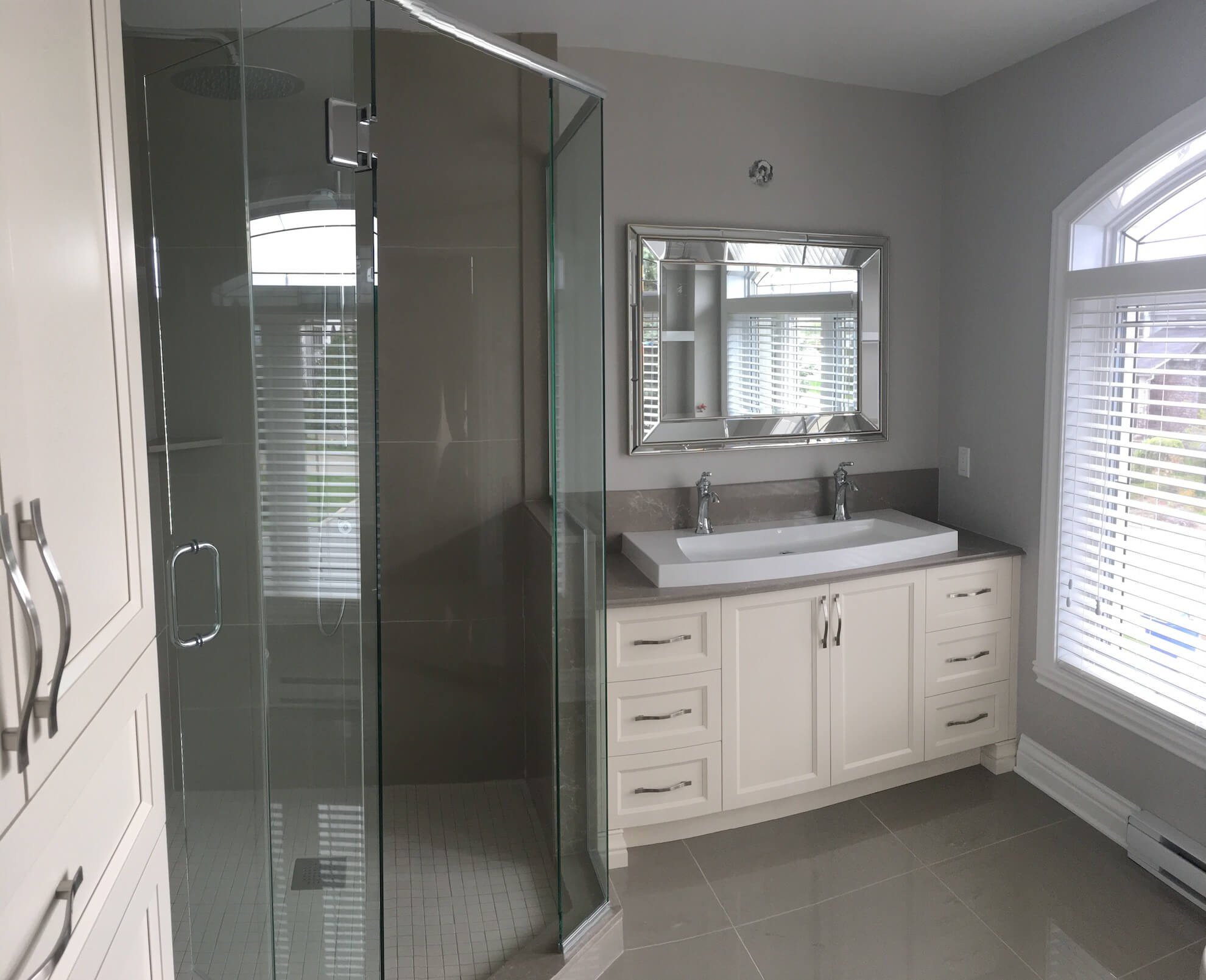 Une salle de bains moderne avec une double vasque blanche, une douche d'angle vitrée, des murs gris et une grande fenêtre avec stores.