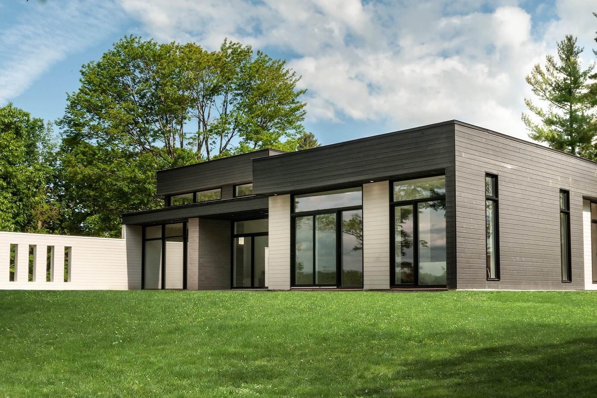 Maison moderne avec murs en verre, extérieur gris et noir, et une pelouse verte, des arbres en arrière-plan.