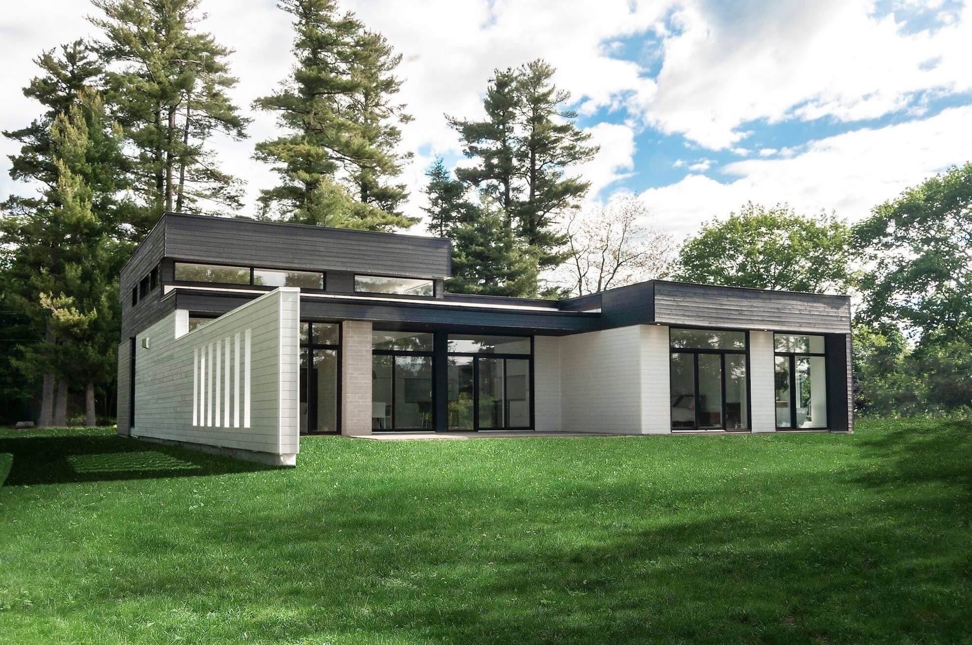 Maison moderne blanche et noire avec de grandes fenêtres, située sur une pelouse verte, entourée d'arbres.