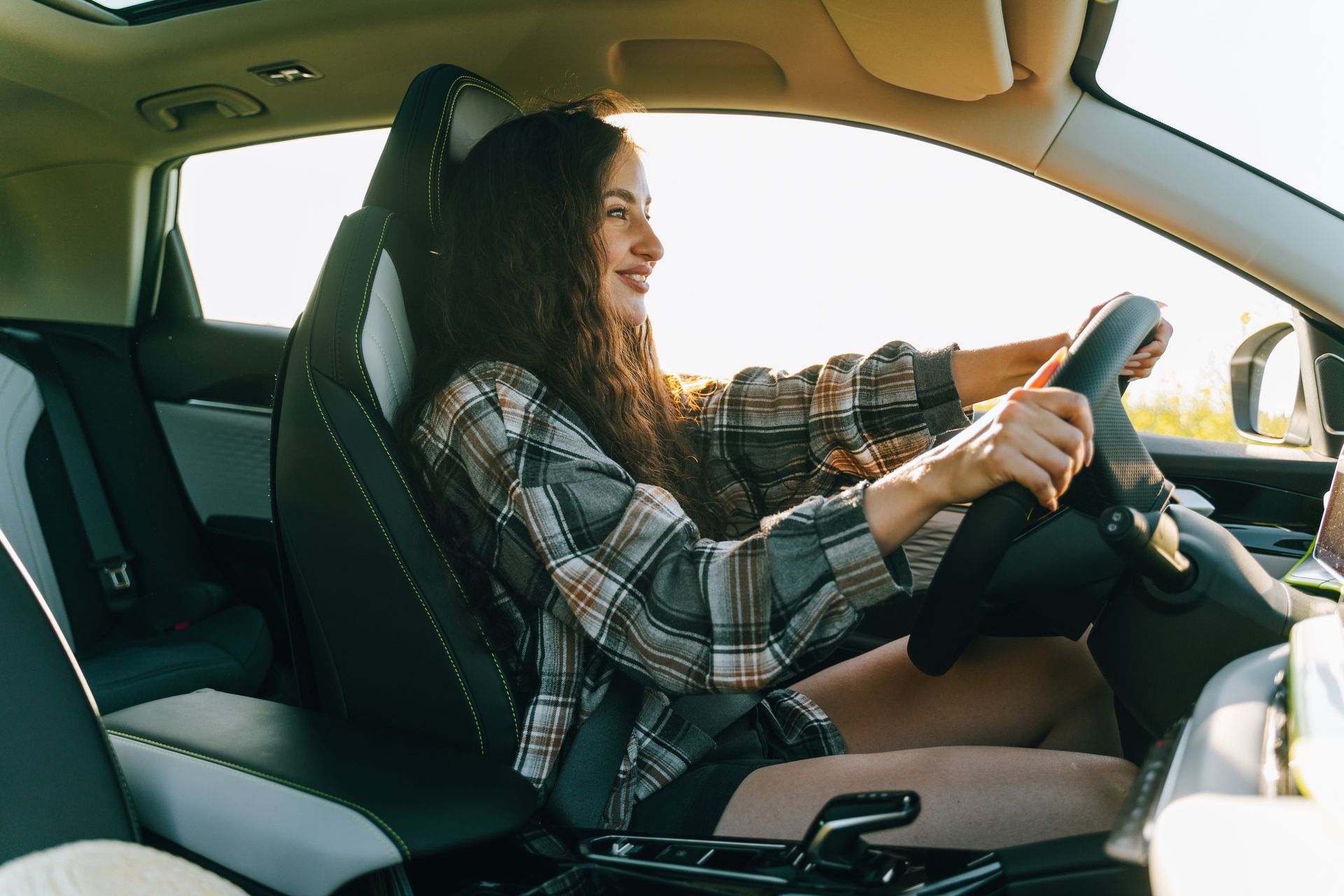 A person with long wavy hair, wearing a plaid shirt, driving a car and smiling while looking ahead.