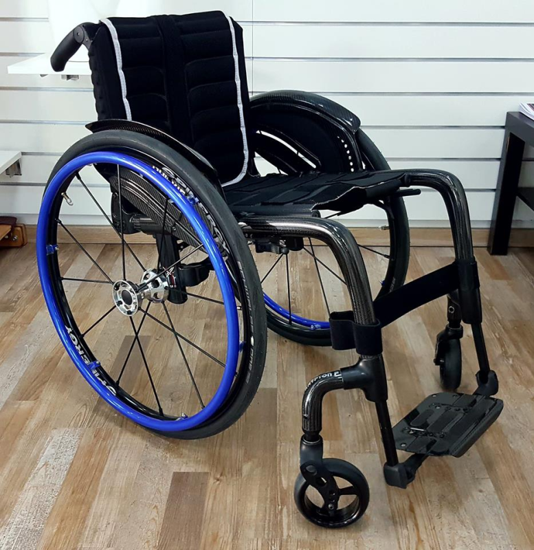 Sedia a rotelle nera con cerchi blu su un pavimento di legno, contro una parete bianca.