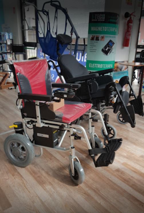 Sedia a rotelle elettrica rossa e nera in un garage, con un caricabatterie e altri dispositivi di mobilità sullo sfondo.