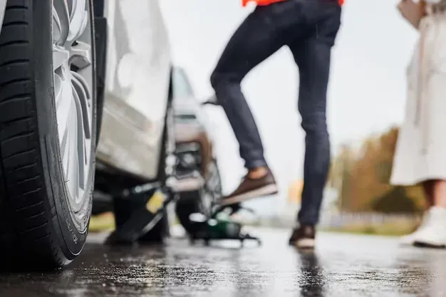 Person changing a car tire on a wet roadside, with a jack and spare wheel nearby