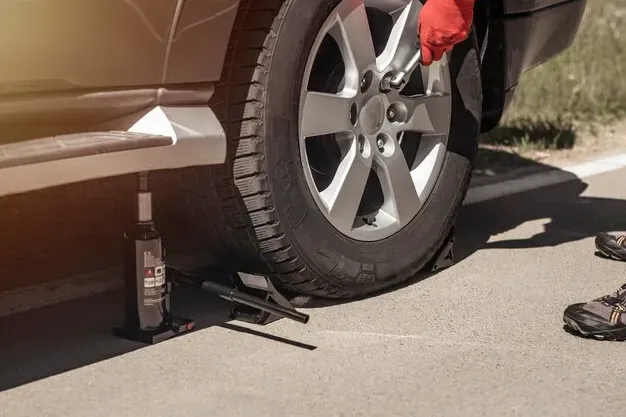 Person changing a flat tire beside a brown car, with a jack and lug wrench on the ground.