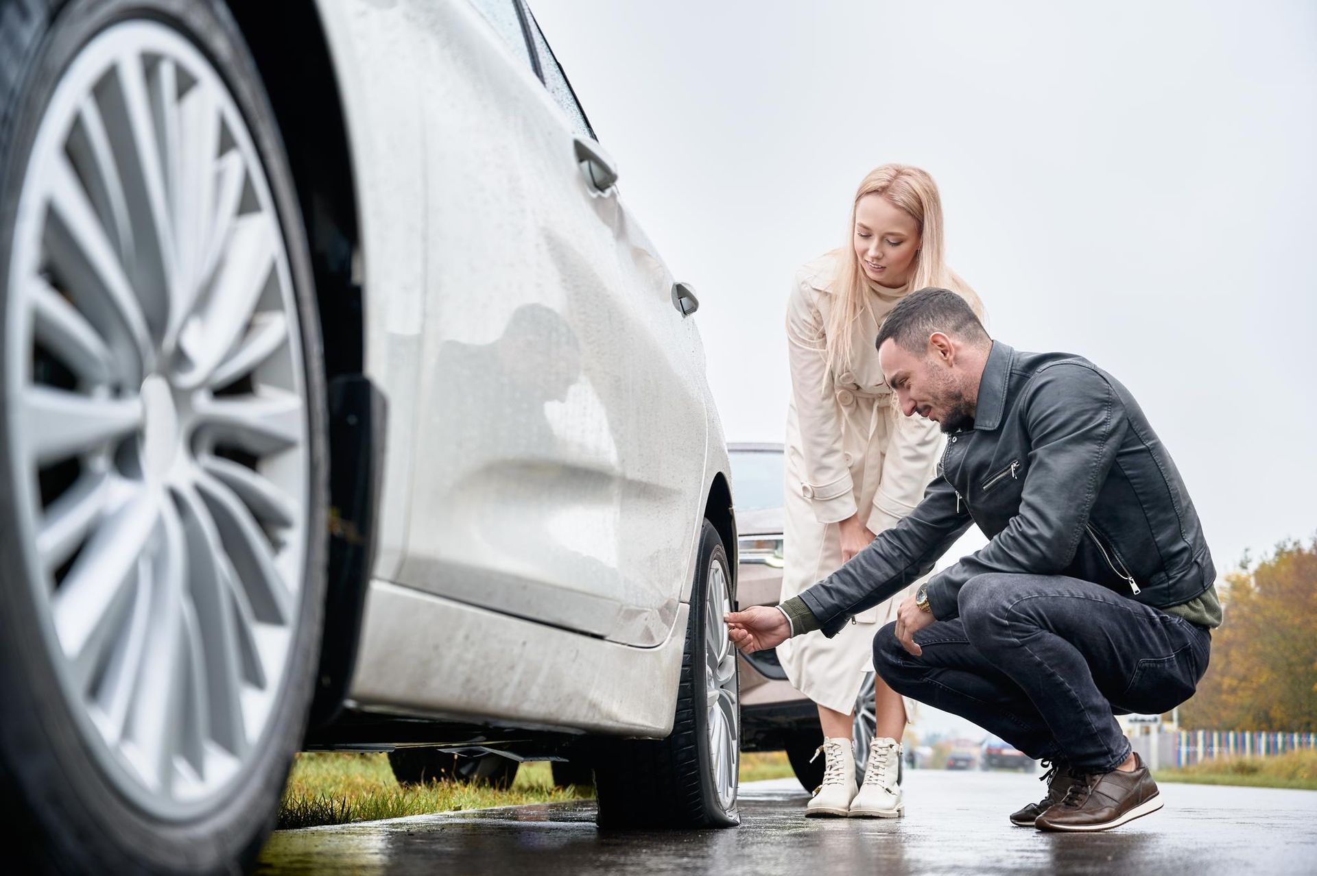 Two people inspect a white car’s flat tire on a wet roadside