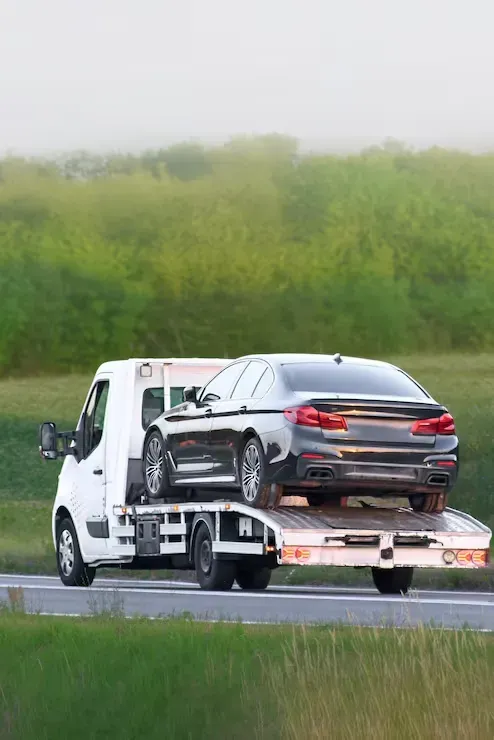 Tow truck hauling a damaged dark sedan on a highway, with green trees in the background