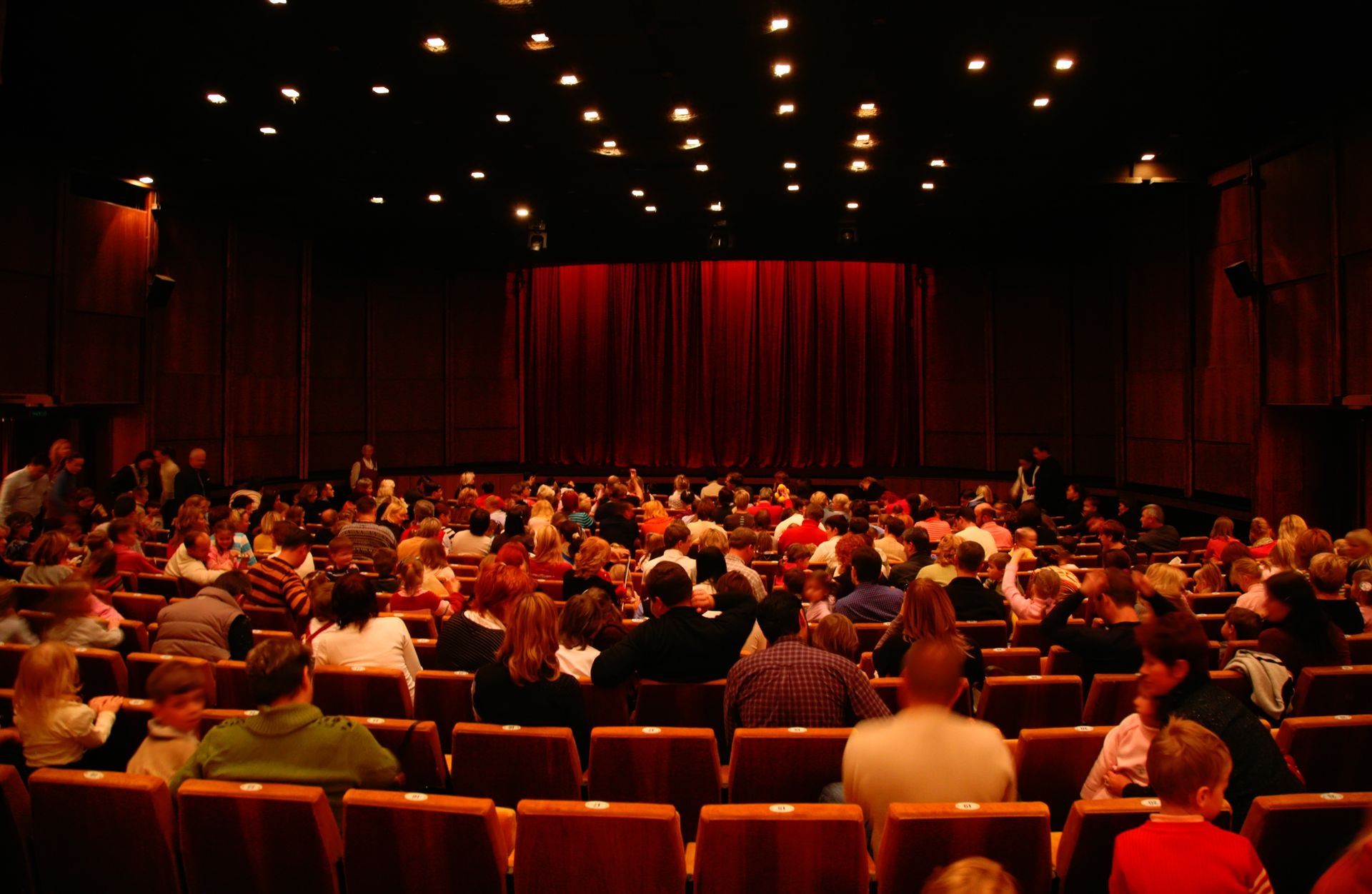 Audience seated in a dark theater facing a red curtain on stage Audience seated in a dark theater facing a red curtain on stage