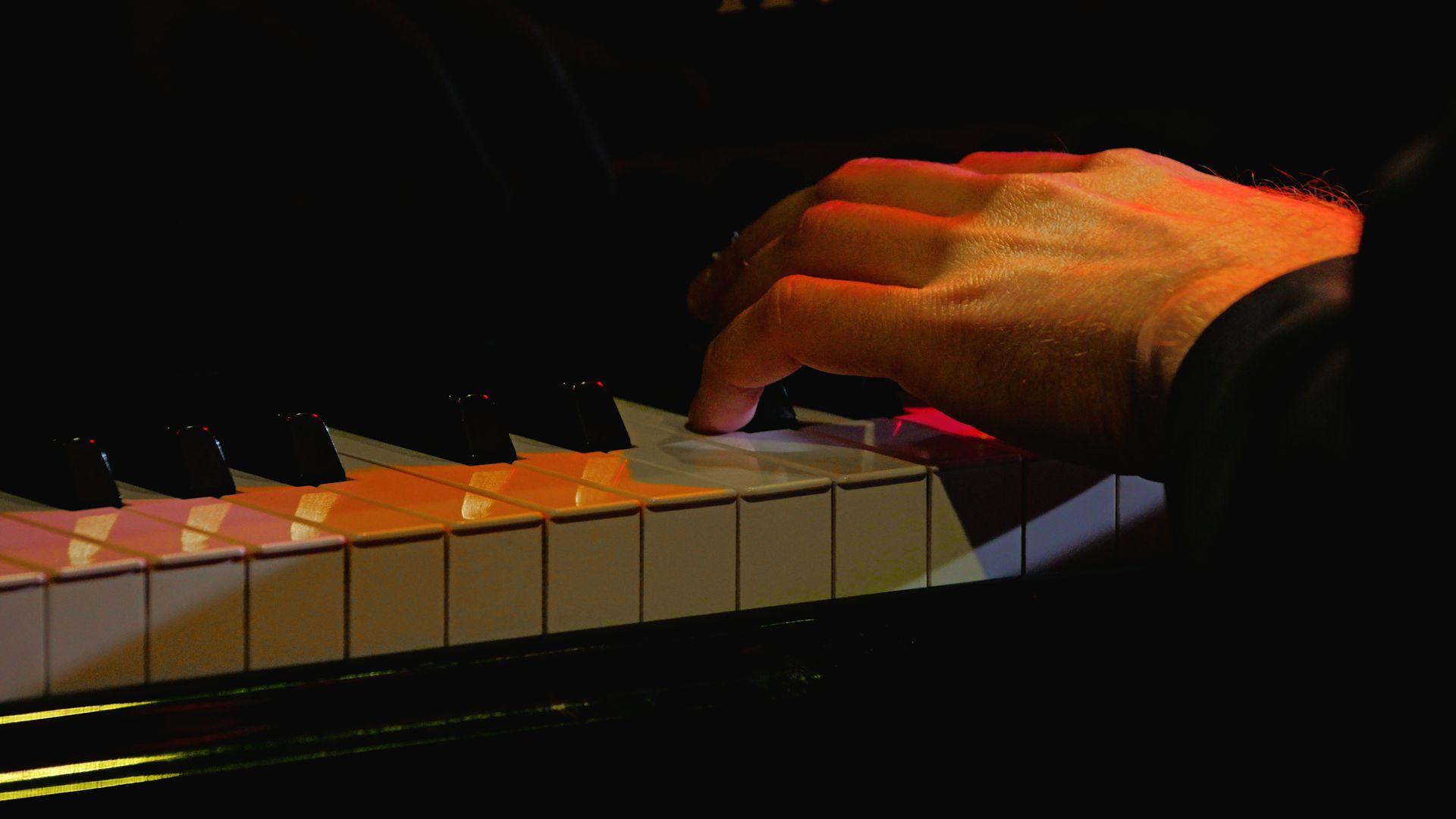 Hand playing a piano keyboard in dim light, with orange highlights on the fingers Hand playing a piano keyboard in dim light, with orange highlights on the fingers