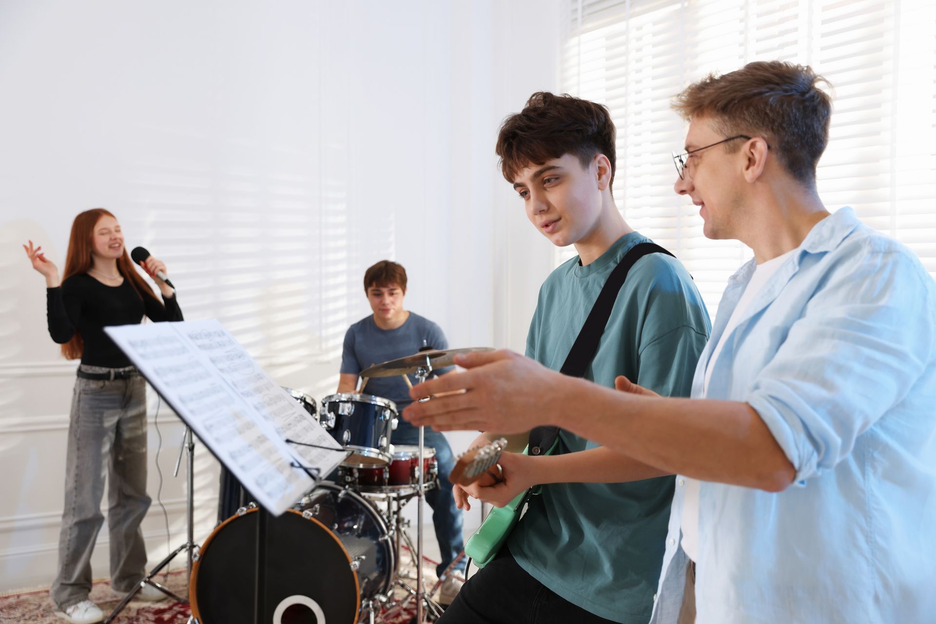 Musicians rehearsing in a bright room, with singer, drummer, and guitarist near a music stand