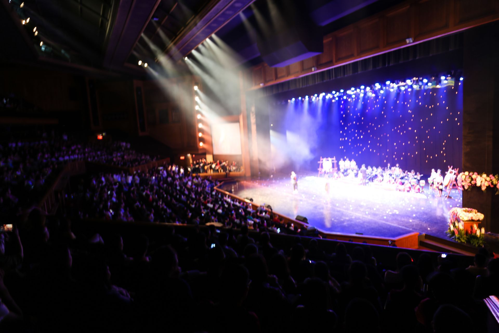 Auditorium with a brightly lit stage, blue backdrop, and a large audience watching a performance Auditorium with a brightly lit stage, blue backdrop, and a large audience watching a performance