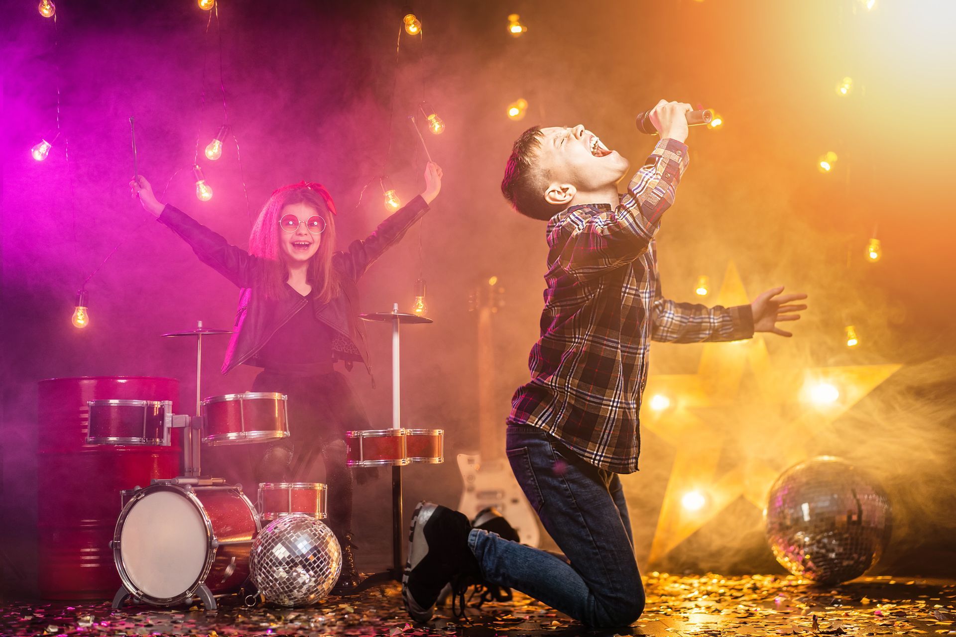 Singer kneels under colorful stage lights while a drummer plays behind him amid smoke and confetti