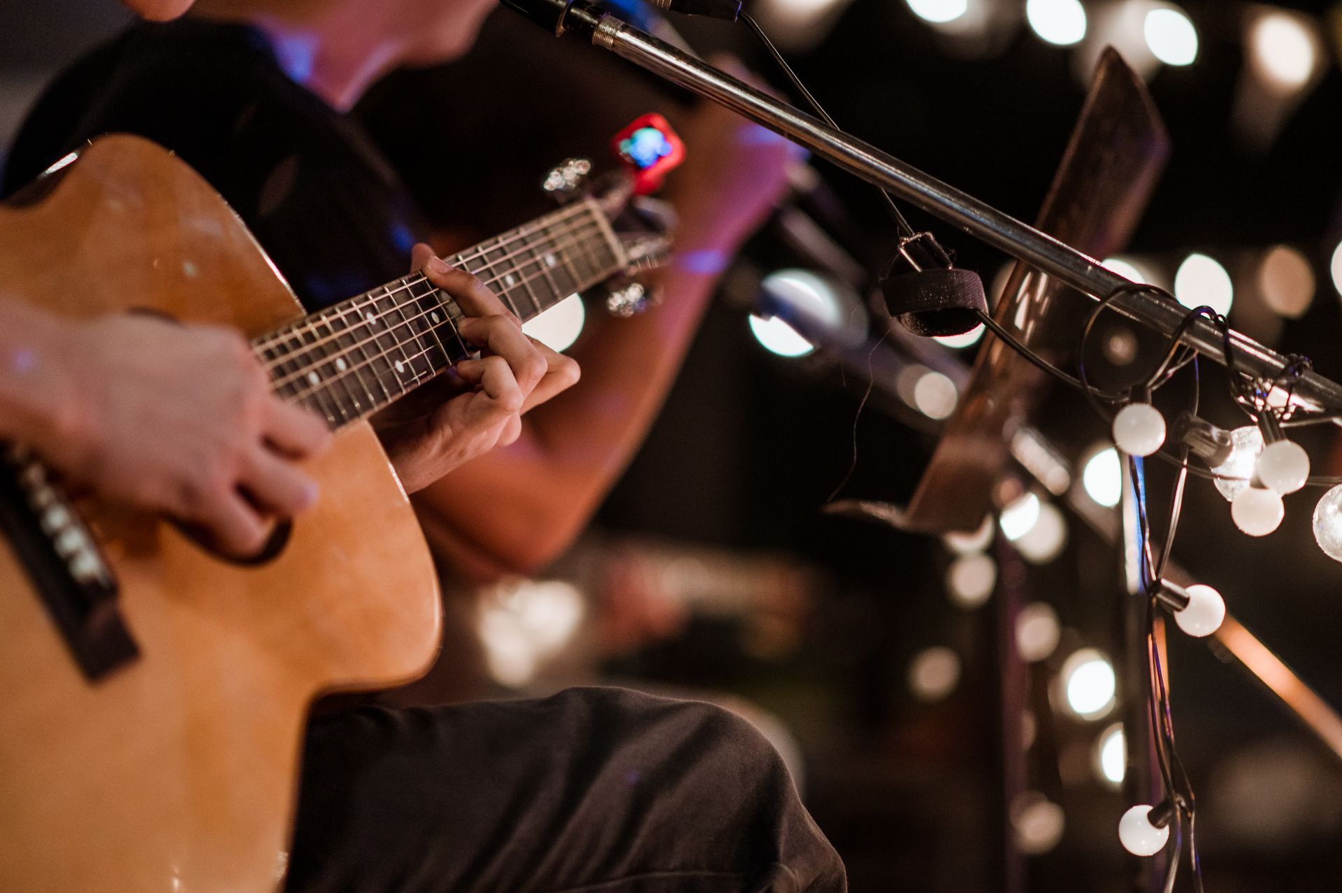 Musician playing acoustic guitar beside warm string lights at a dimly lit performance Musician playing acoustic guitar beside warm string lights at a dimly lit performance