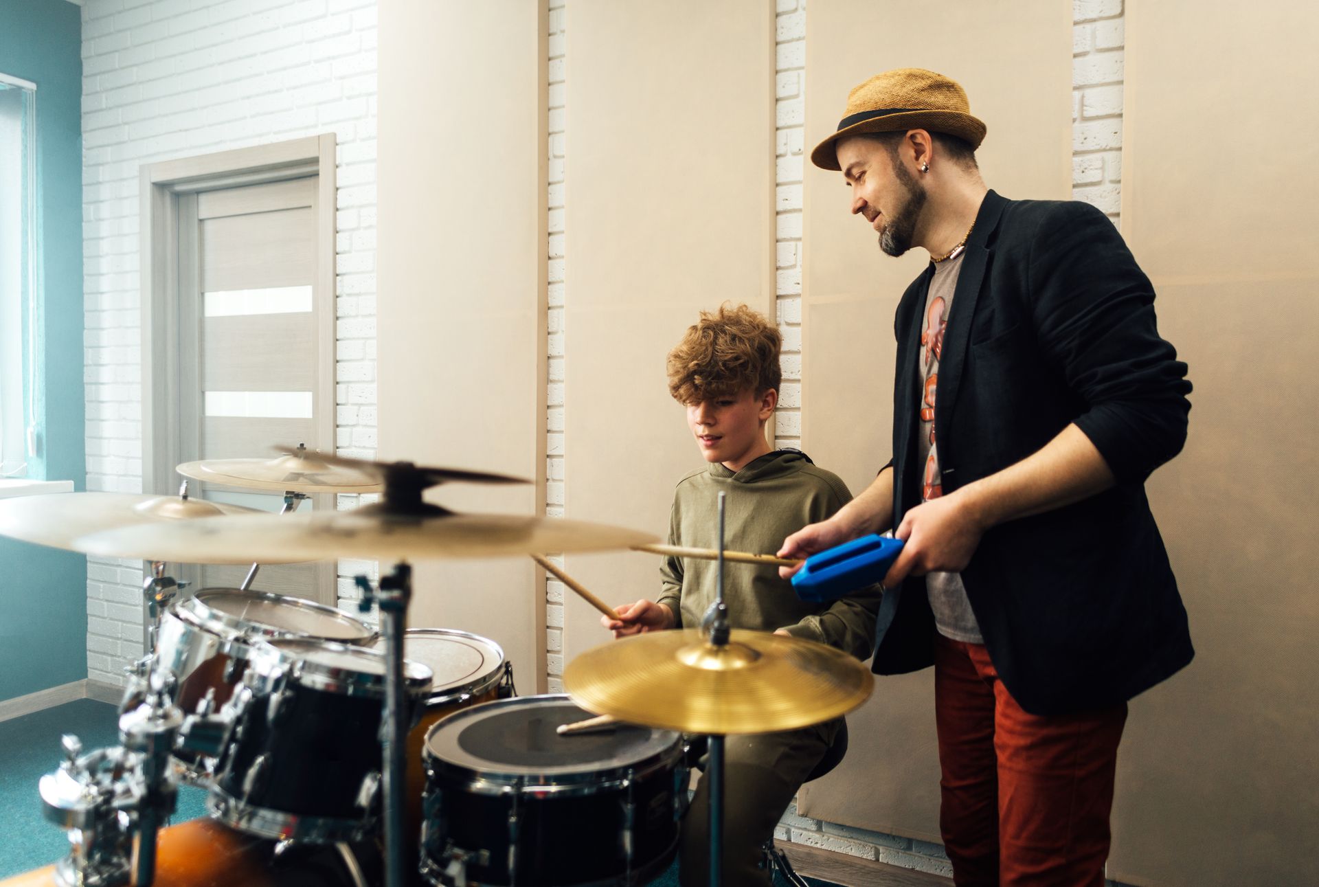 Two people in a music room, one playing drums while another stands beside him holding a blue mallet.