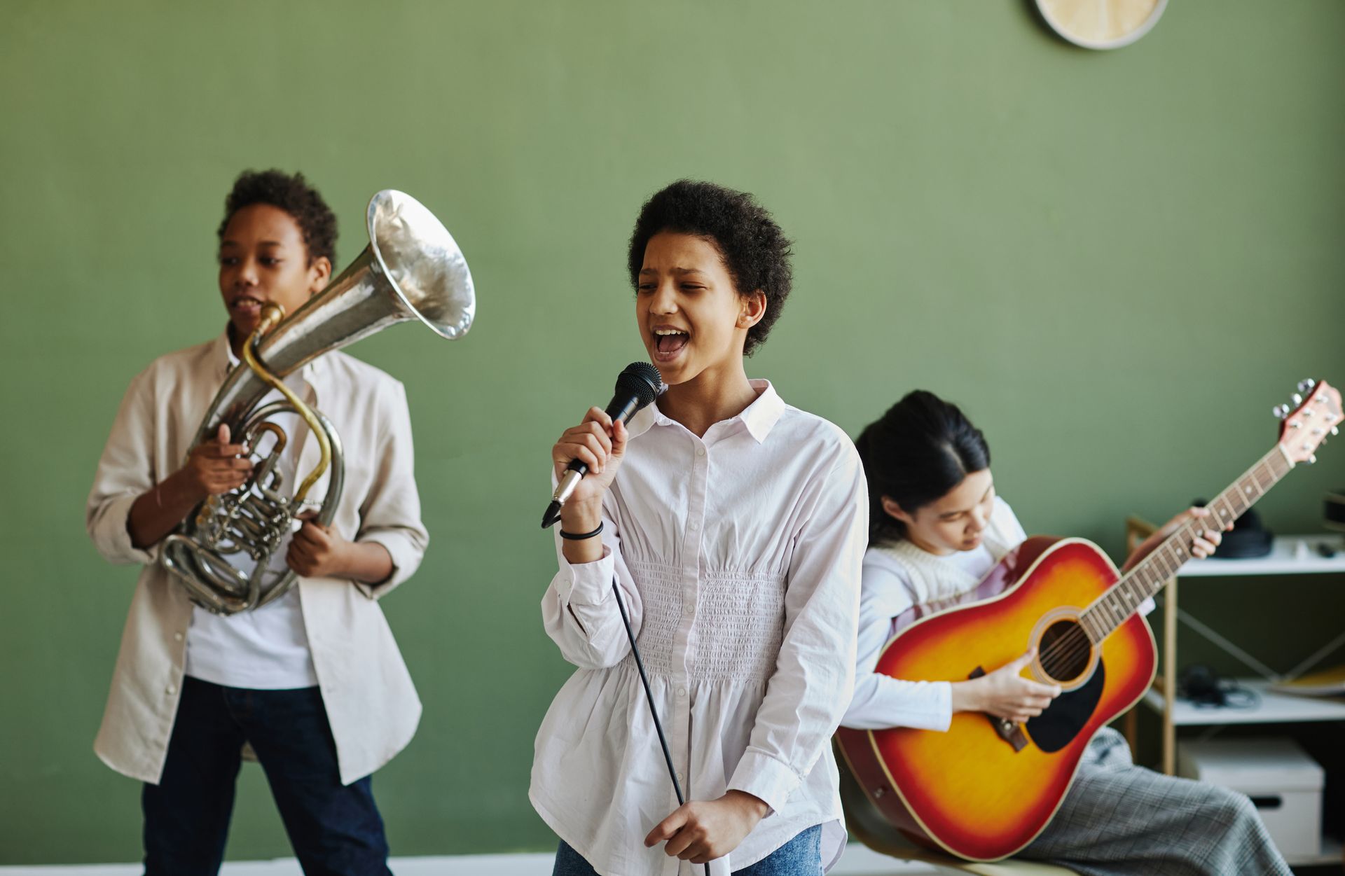 Three children play brass, microphone, and guitar in a classroom with a green wall.