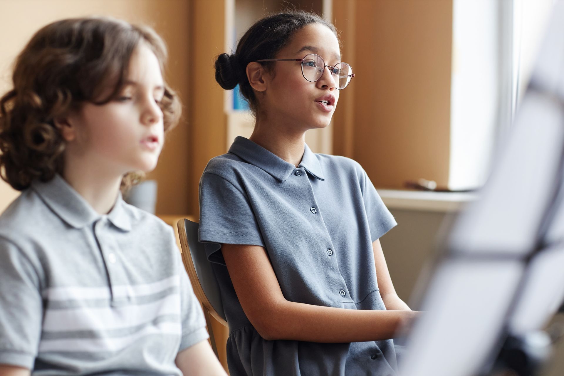 Two children in gray shirts seated in a classroom, one speaking while music stands blur in the foreground Two children in gray shirts seated in a classroom, one speaking while music stands blur in the foreground