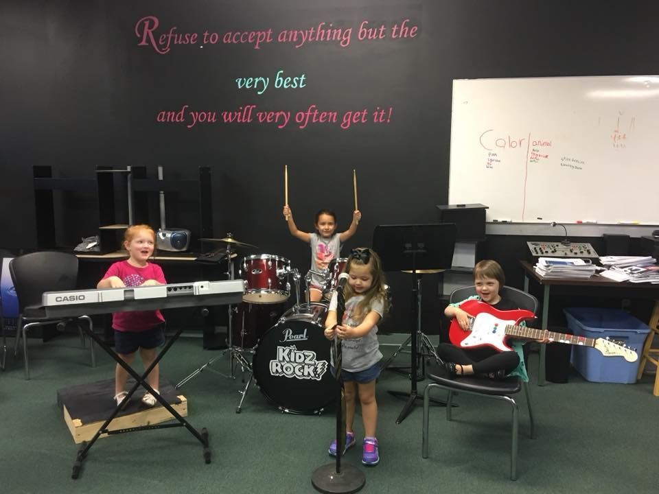 Four musicians performing against a black brick wall, with a singer in a yellow jacket center stage.