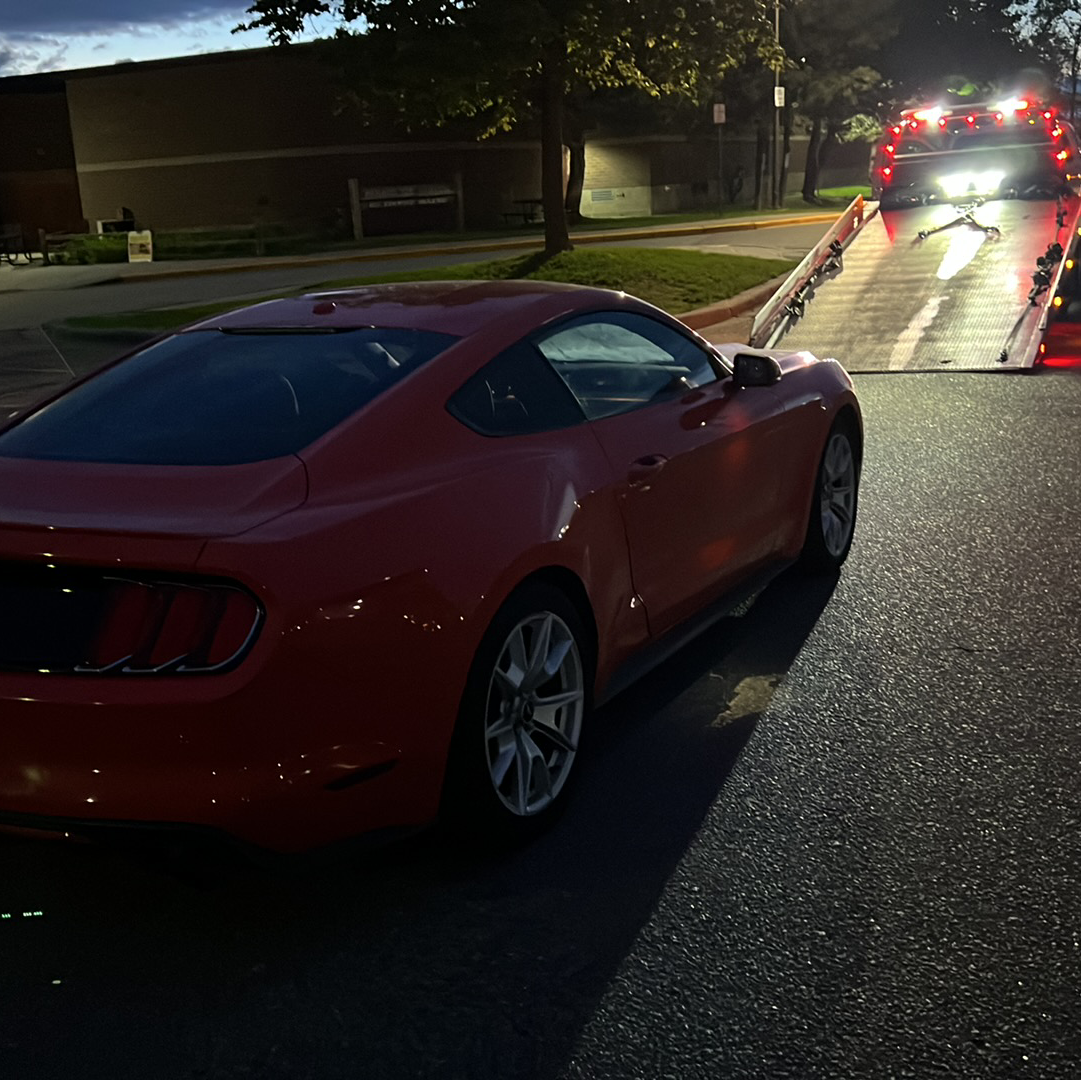 Red sports car on a tow truck at dusk, with emergency lights ahead on a residential street.