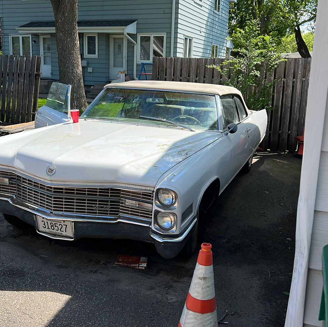 White classic Cadillac parked in a driveway beside a house and fence, with traffic cones in front.