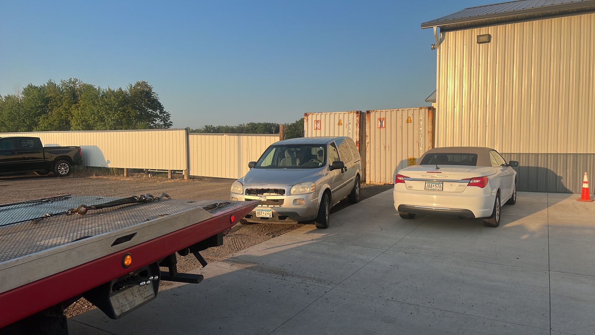 Car lot with a tow truck ramp, tan SUV, white sedan, and a warehouse building at sunset
