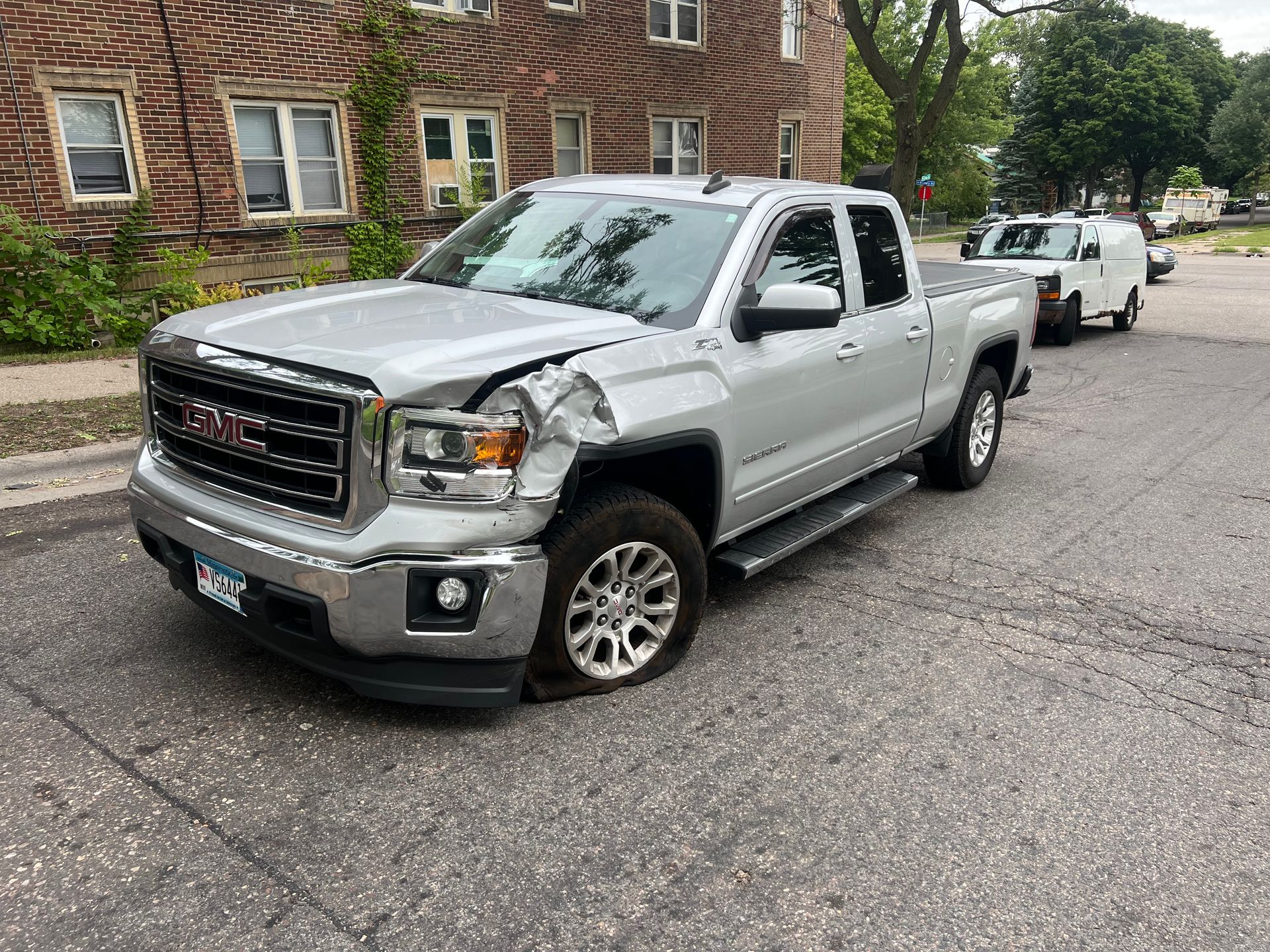 Silver pickup truck with front-end damage parked on a residential street, with a second car behind it