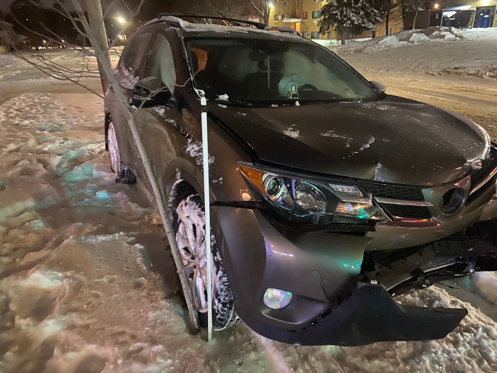 Dark sedan stuck in deep snow at night, with front damage and a snow pole beside it