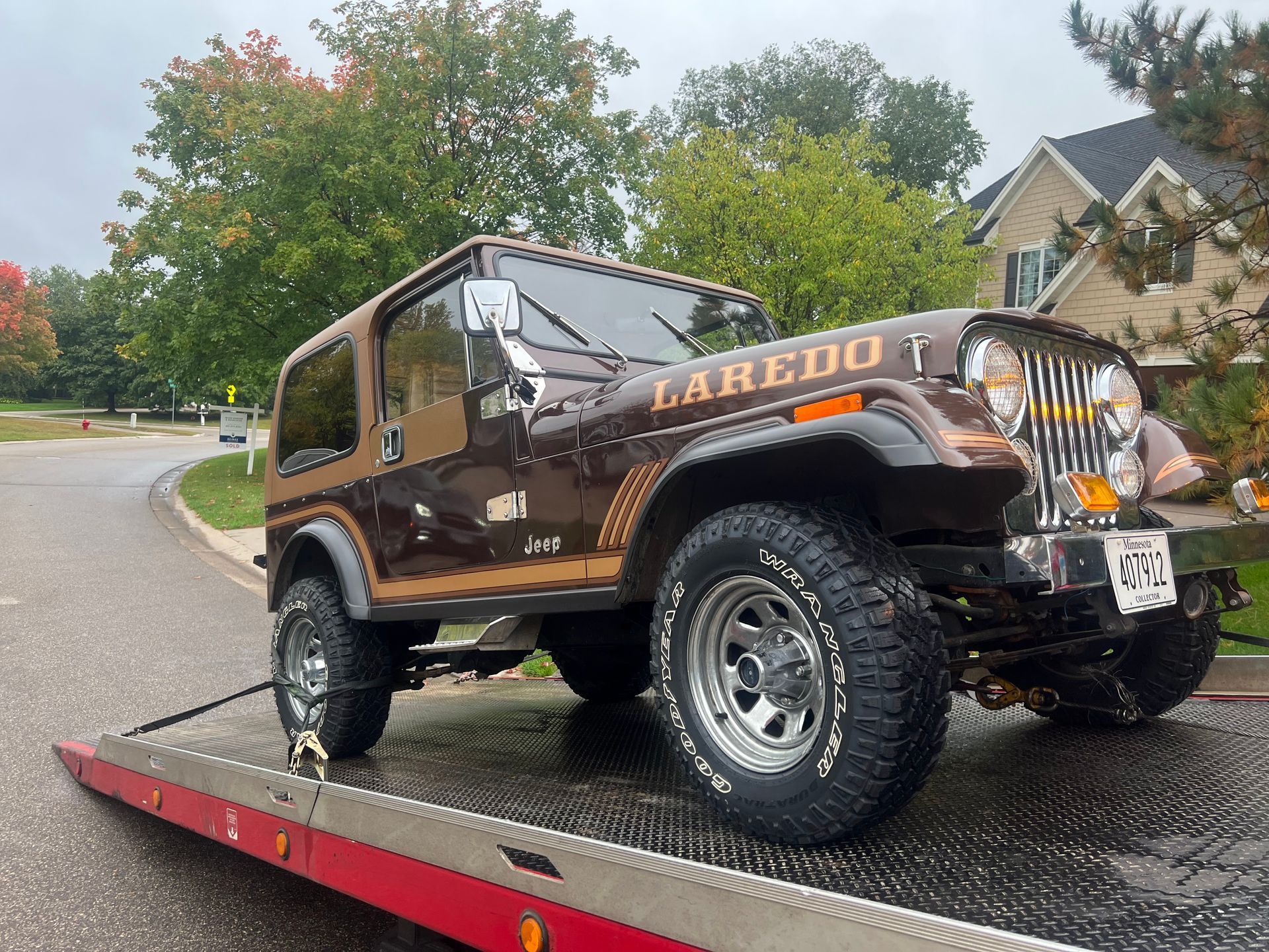 Brown Jeep Wrangler on a red trailer, parked on a wet road in a leafy neighborhood.