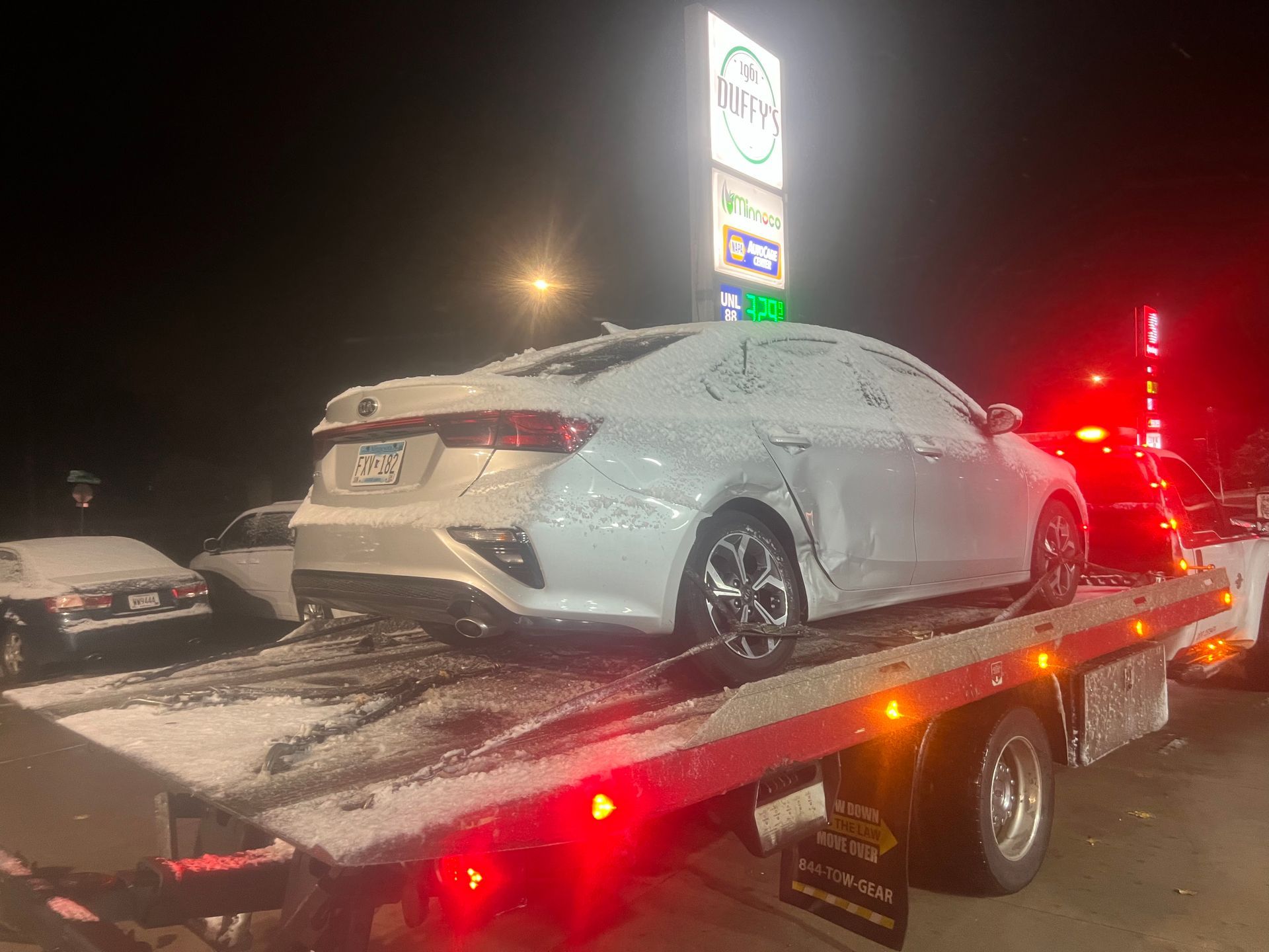 Snow-covered white sedan on a tow truck at night with red lights glowing