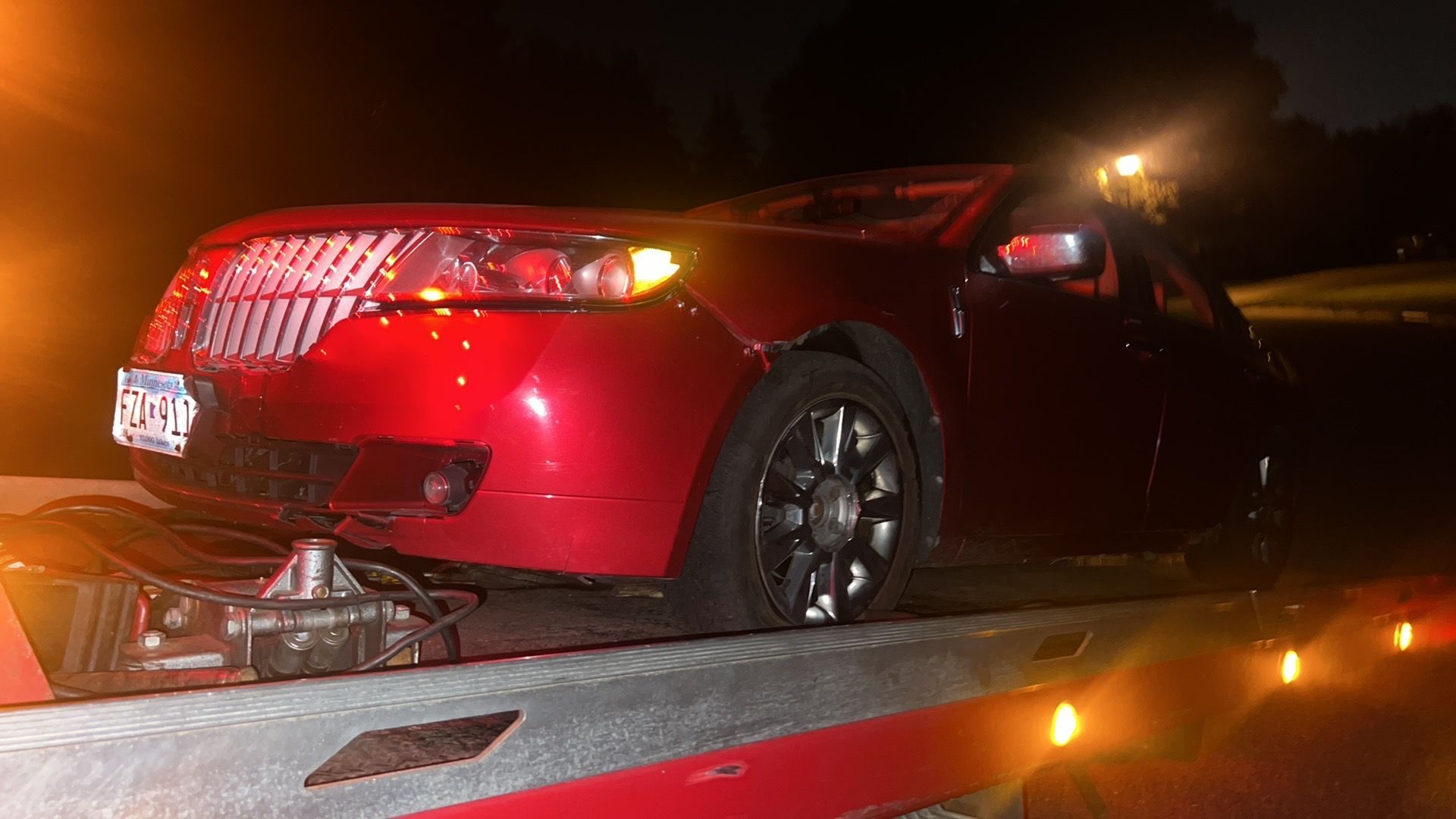 Red sports car on a tow truck at night, rear damaged and lit by flashing lights