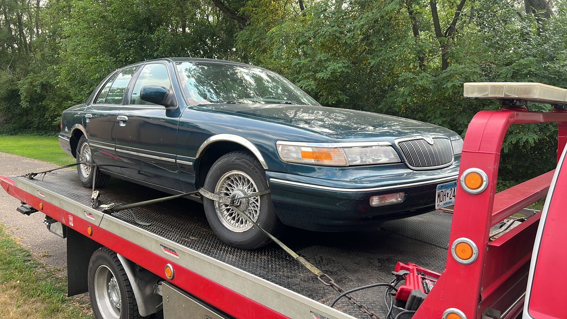 Dark green sedan on a red tow trailer beside a wooded roadside.