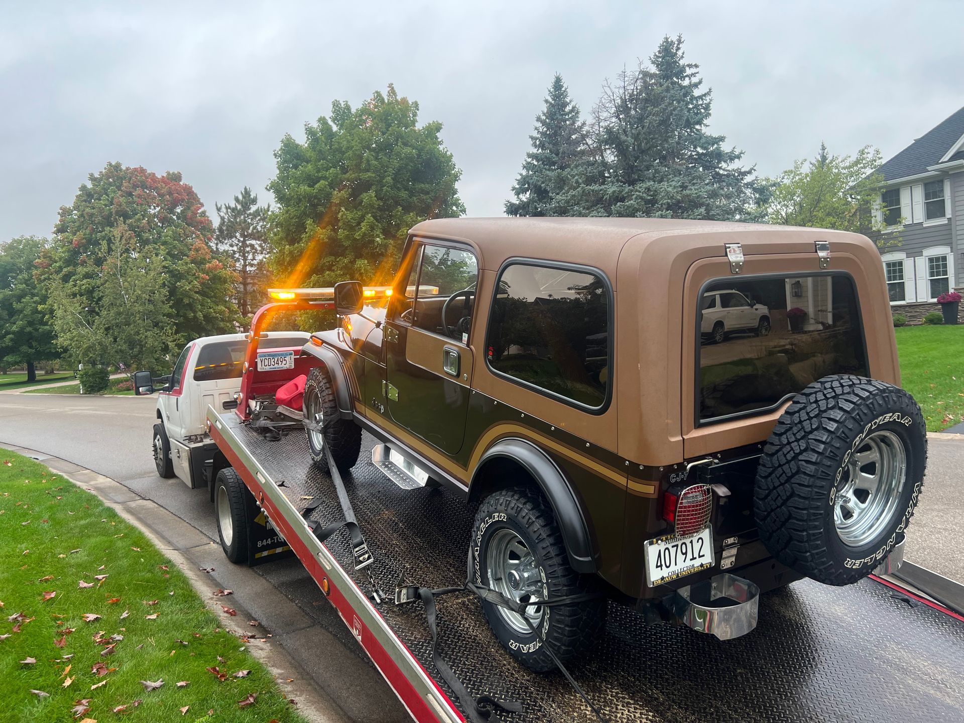 Brown Jeep on a tow truck on a wet suburban street with autumn trees.