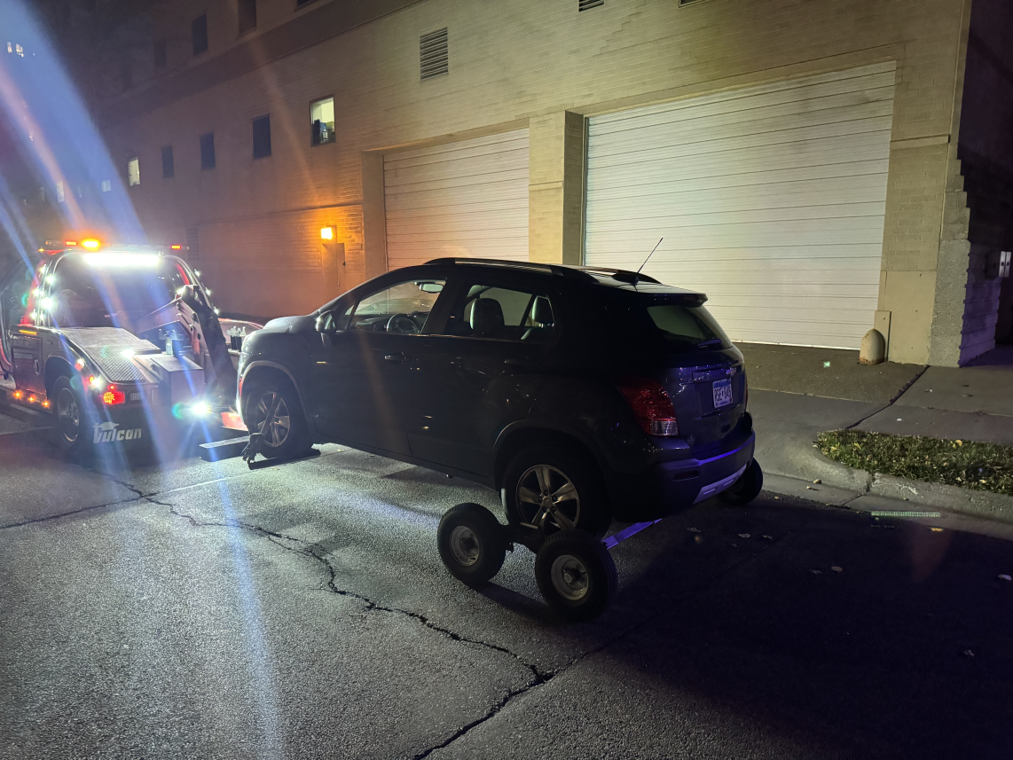 Black SUV on a tow dolly being pulled on a city street at night, with a tow truck’s lights flashing nearby